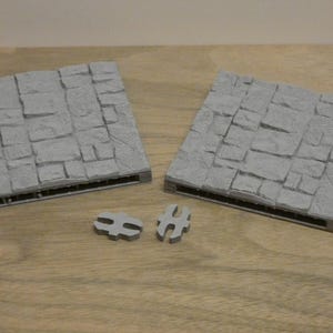 May include: Two grey, square terrain tiles with a cobblestone texture, and two smaller connector pieces. The tiles have a rectangular base with a grid pattern. The items are on a wooden surface.