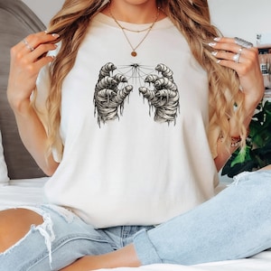 May include: Cream-colored t-shirt featuring a black and white graphic of two wrapped hands reaching towards a central point with connecting lines. The design has a spooky, Halloween theme.