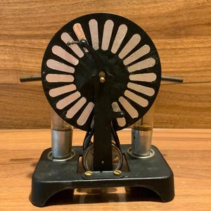 May include: A vintage scientific apparatus featuring a black circular wheel with white radial slots mounted on a black metal base. Two clear glass cylindrical components are positioned vertically on either side of the central support structure.