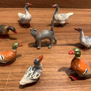 Op de afbeelding: Een verzameling vintage metalen dierenbeeldjes, waaronder eenden, ganzen en een kat. De eenden hebben oranje en groene kleuren, terwijl de ganzen wit en grijs zijn. De kat is grijs met een rode halsband. Alle beeldjes hebben rode poten.