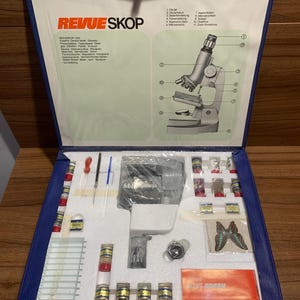 May include: A blue case containing a Revue Skop 1000 microscope kit. The kit includes a microscope, slides, vials, and a butterfly illustration. The case has the Revue Skop logo and text in German.