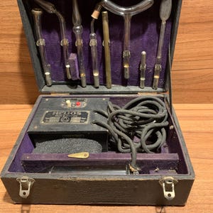 Puede incluir: Un estuche negro vintage abierto, con interior de terciopelo morado. Contiene tubos de vidrio y un dispositivo con la inscripción "HELIOS Made in Germany". También se ven un cable negro y un asa de cuero.