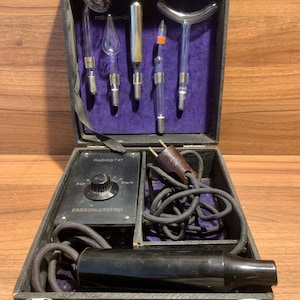Könnte beinhalten: Ein Radiostat Violet Ray Gerät in einem schwarzen Koffer mit violettem Samtfutter. Das Set enthält fünf Glaselektroden in verschiedenen Formen, eine schwarze Steuereinheit mit einem Drehregler mit der Beschriftung Aus und Stark sowie einen Handgriff.
