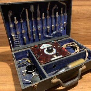 Puede incluir: Una antigua máquina de alta frecuencia Zenith en una caja de madera gris oscuro. La caja está forrada con terciopelo azul y contiene tubos de vidrio, un panel de control y gafas de sol redondas. El asa es de metal curvado y texturizado.