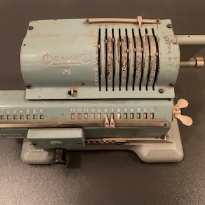 Op de afbeelding: Vintage lichtblauwe mechanische rekenmachine met meerdere nummerwielen en een slinger. De rekenmachine heeft een reeks nummerwielen en hendels, met het woord "Феликс" zichtbaar aan de voorkant.