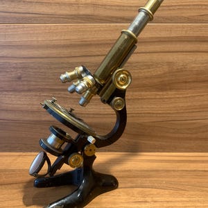 May include: An antique microscope with a black base and brass components. The microscope has multiple lenses and adjustment knobs. The eyepiece is silver-colored, and the overall design suggests a vintage scientific instrument.