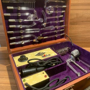 Puede incluir: Kit de dispositivos médicos antiguos en una caja de madera. La caja abierta revela aplicadores de vidrio, un panel de control con la palabra "Triumph" y otros componentes. El interior está forrado con terciopelo morado.