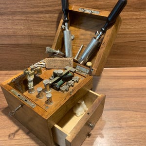 Peut inclure: Une boîte en bois contenant un mécanisme de télégraphe avec des composants métalliques, deux outils à manche noir dans le couvercle et un petit tiroir ouvert. L'appareil présente divers boutons, vis en laiton et argent, et une pièce cylindrique verte.