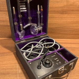 May include: A vintage Juwel 4523 high frequency violet ray device in a black carrying case with purple velvet lining. The set includes a black control unit with a dial, a power cord, and four glass electrodes of various shapes for skin treatment.