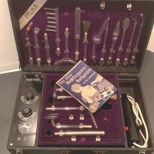 Puede incluir: Un maletín negro vintage abierto, con un interior de terciopelo morado, que contiene varios instrumentos médicos de vidrio. El maletín incluye un panel de control y un folleto titulado "Bau eines Hochfrequenz-Heilgeräts". La marca "FELMA" es visible.