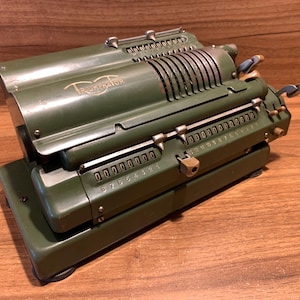 May include: An olive green Triumphator mechanical calculator. The vintage machine has multiple rows of number dials, levers, and a metal casing. The word "Triumphator" is printed on the top. The calculator sits on a wooden surface.