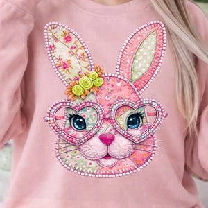 Peut inclure: Sweat-shirt rose clair avec un motif de visage de lapin coloré. Le lapin porte des lunettes en forme de cœur, des oreilles à motifs floraux et un nez rose. Le motif comprend une couronne florale et des détails cousus, parfait pour Pâques.