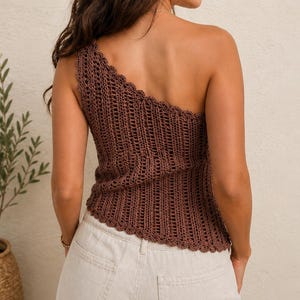 May include: A brown crochet one-shoulder top featuring a vertical ribbed pattern and a scalloped edge along the neckline and hem. The garment is paired with light-colored denim jeans that have visible back pockets and a button closure.