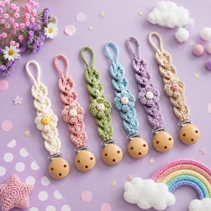 May include: Six crocheted pacifier clips in cream, pink, green, light blue, purple, and beige. Each features a braided design with a central flower motif, a loop at the top, and a round wooden clip with three holes at the bottom for attachment.