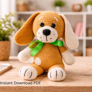 May include: A handmade crochet dog toy in shades of tan, cream, and black. The dog has large floppy ears, a black nose, and a green ribbon around its neck. The toy is sitting on a wooden surface. The text "Instant Download PDF" is at the bottom.