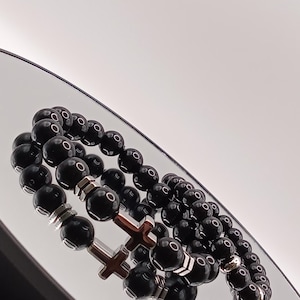 Puede incluir: Dos pulseras de cuentas con cuentas redondas negras. Una pulsera presenta detalles metálicos plateados y dos dijes de cruz de madera. La otra pulsera tiene detalles metálicos plateados. Las pulseras se exhiben sobre una superficie reflectante.