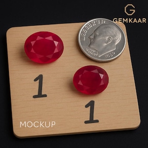 Op de afbeelding: Twee gefacetteerde, ovale, rode edelstenen worden getoond op een houten oppervlak met een munt voor schaalvergelijking. Op het houten oppervlak staan het nummer "1" en het woord "MOCKUP" afgedrukt. Het Gemkaar-logo staat rechtsboven.