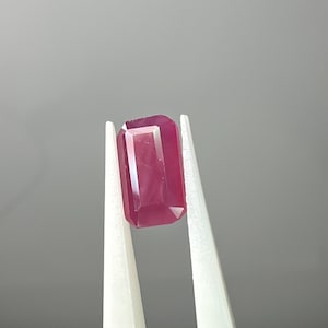 Puede incluir: Un rubí rectangular facetado sostenido por pinzas blancas. La gema es de color rojo rosado intenso y tiene una superficie clara y reflectante. La piedra preciosa mide aproximadamente 1,9 cm de largo.