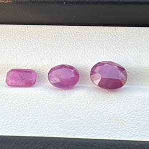 Könnte beinhalten: Drei polierte Rubin-Edelsteine in verschiedenen Formen und Größen. Die Edelsteine haben eine tiefrosa-rote Farbe und werden in einer weißen Box präsentiert. Die Formen umfassen ein Rechteck, ein Oval und ein größeres Oval.