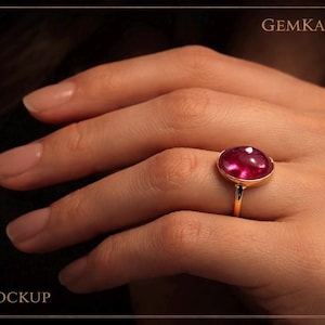 May include: A gold ring with a large, oval, ruby-colored gemstone. The ring is set on a slender gold band. The image includes the text "GEMKAAR" and "MOCKUP".