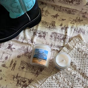 Puede incluir: Una bota vaquera de cuero negro y turquesa junto a dos frascos de vidrio con velas de cera blanca. Una vela tiene una etiqueta que dice literally Rebel Blue Ranch con una ilustración de paisaje desértico sobre una tela beige con escenas.