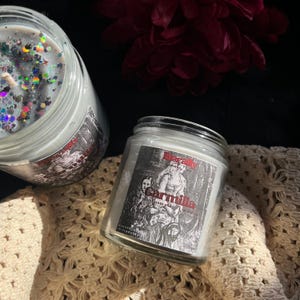 Può includere: Due candele in barattolo di vetro con cera bianca ed etichette con un'illustrazione vintage e la parola "Carmilla" in rosso. Una candela ha glitter. Le candele sono su una superficie color crema a trama aperta, con un fiore rosso scuro sullo sfondo.
