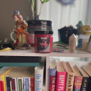 Könnte beinhalten: Ein zylindrisches Glasgefäß mit schwarzem Deckel, das rotes Wachs enthält. Das Etikett zeigt den Text "literally" und "Nina" in Fettdruck sowie eine Grafik von Händen. Das Glas steht auf einem weißen Regal zwischen verschiedenen Büchern.