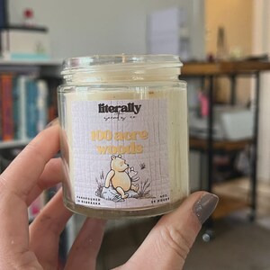 May include: A clear glass jar candle with a cream-colored wax fill. The label features the text "literally scents co", "100 acre woods", "handpoured in nebraska", "4oz.", and "25 hours" alongside an illustration of a yellow bear sitting on a rock.