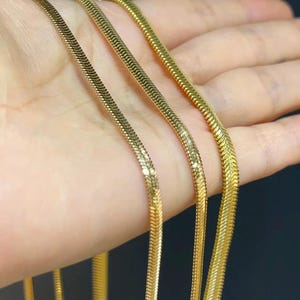 May include: Three gold-toned snake chains featuring a flat, interlocking herringbone pattern with a polished, reflective finish. Each chain is flexible and displays a consistent, smooth texture throughout its length, ending in a small metal clasp.
