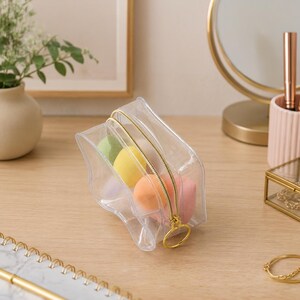 K&ouml;nnte beinhalten: Eine transparente Kosmetiktasche mit goldenem Rei&szlig;verschluss, gef&uuml;llt mit bunten Make-up-Schw&auml;mmen. Die Tasche liegt auf einer Holzoberfl&auml;che, mit einem goldgerahmten Spiegel und anderem Sch&ouml;nheitszubeh&ouml;r im Hintergrund.