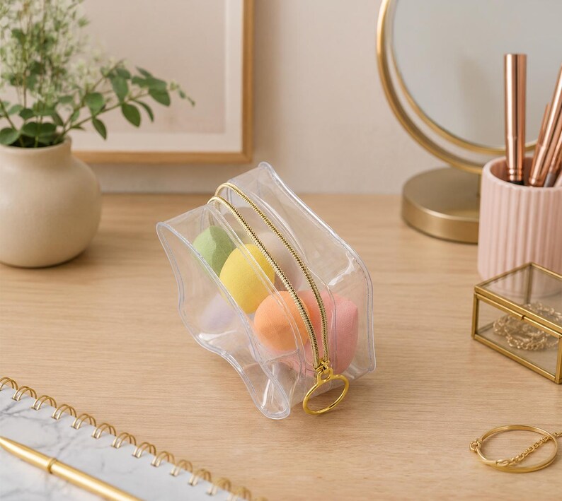 K&ouml;nnte beinhalten: Eine transparente Kosmetiktasche mit goldenem Rei&szlig;verschluss, gef&uuml;llt mit bunten Make-up-Schw&auml;mmen. Die Tasche liegt auf einer Holzoberfl&auml;che, mit einem goldgerahmten Spiegel und anderem Sch&ouml;nheitszubeh&ouml;r im Hintergrund.