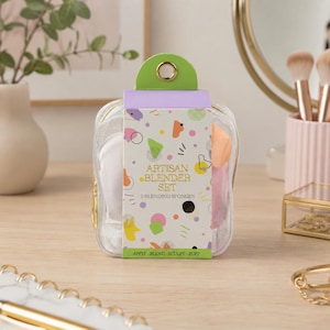 K&ouml;nnte beinhalten: Eine transparente Kosmetiktasche mit einem gr&uuml;nen und goldenen Griff enth&auml;lt ein Set aus f&uuml;nf Make-up-Schw&auml;mmen. Die Verpackung tr&auml;gt den Text "ARTISAN BLENDER SET" und bunte geometrische Formen. Auf der Unterseite der Verpackung steht "APPLY - BLEND - SCULPT - BUFF."