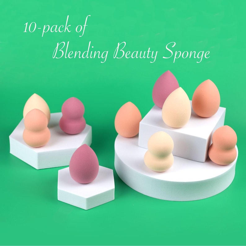 May include: A 10-pack of Blending Beauty Sponge items in various shades of pink, peach, and cream. The sponges are shaped like teardrops or hourglasses and are displayed on white geometric platforms against a solid green background.
