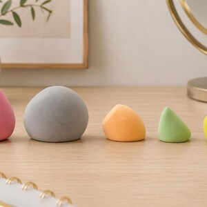 K&ouml;nnte beinhalten: F&uuml;nf farbige Make-up-Schw&auml;mme in verschiedenen Formen und Gr&ouml;&szlig;en sind auf einer Holzoberfl&auml;che angeordnet. Die Schw&auml;mme sind rosa, grau, orange, gr&uuml;n und gelb. Der Hintergrund umfasst eine Vase mit Gr&uuml;n, einen Spiegel und Make-up-Pinsel.
