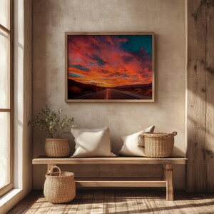 Op de afbeelding: Een ingelijste print toont een weg die leidt naar een zonsondergang met rode, oranje en turquoise kleuren. Het kunstwerk hangt boven een houten bank met geweven manden en kussens, wat bijdraagt aan een gezellig interieur.