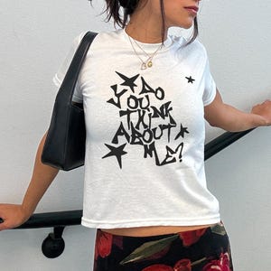 Può includere: Una t-shirt bianca a maniche corte con testo grafico nero che recita "DO YOU THINK ABOUT ME?" circondato da varie forme di stelle. La maglietta è abbinata a una borsa a tracolla nera e a una gonna nera con motivo floreale rosso e verde.