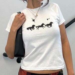 May include: White crew neck t-shirt with a black graphic of three running horses, stars, and a celestial design. The model is wearing black sunglasses and a black purse. The shirt is short-sleeved.