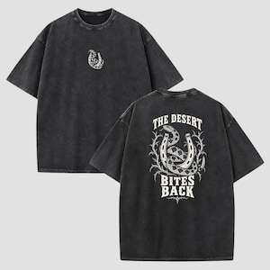 Può includere: Una t-shirt a maniche corte grigio antracite con finitura slavata. La parte anteriore presenta una piccola grafica bianca di un serpente avvolto attorno a un ferro di cavallo. Il retro mostra una grande grafica bianca di un serpente, ferro di cavallo e spine con il testo THE DESERT BITES BACK.