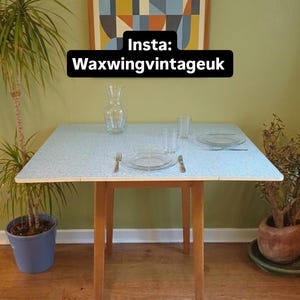 May include: A retro square table with a light blue speckled surface and wooden legs. The table is set with glassware, plates, and cutlery. A framed abstract artwork hangs on the wall. The text "Insta: Waxwingvintageuk" is visible.