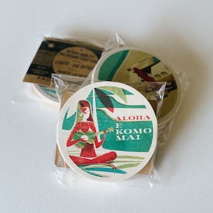 May include: A collection of round coasters with a vintage Hawaiian theme. One coaster features a woman playing a ukulele with the text "Aloha E Komo Mai". The coasters are packaged in clear plastic.