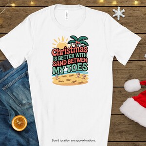May include: White t-shirt with a colorful graphic that reads "Christmas is better with sand between my toes." The design includes a sun, palm trees, and footprints on a beach. The shirt is displayed on a wooden surface.