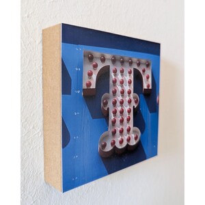 Könnte beinhalten: Ein Holzblock mit einem großen, beleuchteten Buchstaben "T". Der Buchstabe ist weiß mit roten Glühbirnen, vor einem blauen Hintergrund mit einem dezenten Muster. Die Seiten des Blocks sind naturfarbenes Holz.