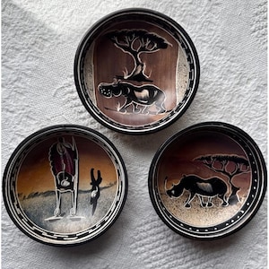 May include: Three round bowls with black rims and white geometric border patterns. The interior of each bowl features a brown and black painted scene: one with a hippopotamus and tree, one with a rhinoceros and tree, and one with a standing figure.