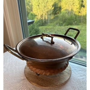 May include: A round, hammered copper serving dish with a matching lid, featuring two brass side handles and a central brass lid knob. The dish rests on a circular, carved wooden base with a scalloped edge, providing a stable foundation.