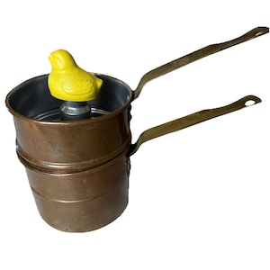 May include: A copper-colored double boiler with two long brass handles. A yellow bird-shaped knob sits atop the central post. The pot has a vintage aesthetic, suggesting it is an antique or a retro kitchen tool.