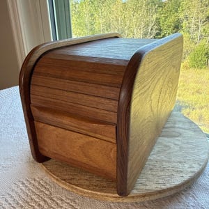 May include: A wooden bread box featuring a curved roll-top lid and a rectangular base. The container is constructed from two distinct tones of wood, with a darker, rich brown finish on the sides and a lighter, natural wood grain on the top.