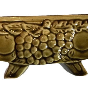 May include: An olive-green ceramic planter with a rectangular shape and three small feet. The planter features a raised relief pattern of grapes and apples. The design is repeated around the exterior of the planter.