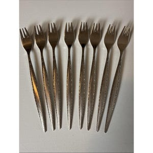 May include: A set of eight stainless steel cocktail forks arranged in a row. Each fork features three tines and a long, slender handle with a textured, brushed metal finish along the edges and a smooth, polished center section.