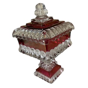 May include: A clear glass candy dish with a ruby red interior and accents. The dish has a square shape with a decorative lid and sits on a pedestal base. The glass is cut with a diamond pattern.