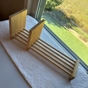 May include: A light-colored wooden rack featuring two vertical rectangular end panels connected by four horizontal cylindrical rods. The structure is designed with a sliding mechanism, allowing the two vertical panels to be adjusted along the rods.